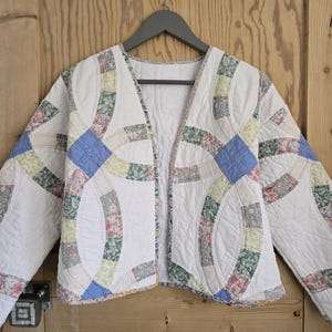 May include: A quilted jacket featuring a white background with a double wedding ring pattern. The design incorporates various floral fabrics in shades of blue, green, yellow, and pink. The jacket has long sleeves, an open front, and a collarless neckline.