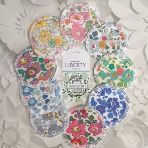 May include: A set of 10 reusable cotton rounds made with Liberty Fabrics. The rounds are circular and feature a variety of floral patterns in shades of pink, blue, green, and purple. The rounds are surrounded by white paper flowers.
