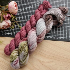 May include: Two twisted hanks of yarn. One hank is a solid deep rose pink. The second hank features a variegated colour pattern with shades of light grey, soft lavender, and patches of muted olive green, creating a multi-tonal appearance.