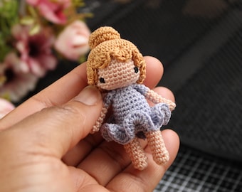 May include: A handmade crochet doll with a light brown bun and hair, wearing a lavender dress. The doll has small black eyes and is made with a detailed crochet stitch. The doll is a miniature size and is held in a hand.