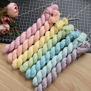 May include: A set of seven twisted yarn skeins arranged in a diagonal gradient pattern, transitioning from soft pink and peach to yellow, light green, light blue, and lavender. Each skein is tightly wound and features a smooth, uniform texture.