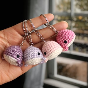 May include: Four crocheted dolphin keyrings in shades of pink, purple, and white. Each keyring has black eyes and a silver key ring.