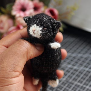 May include: A miniature knitted cat, predominantly black with white markings on its face, paws, and chest. The cat has a small pink nose. The figurine is held in a hand. The background features blurred pink flowers.