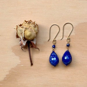 May include: A pair of gold earrings with teardrop-shaped lapis lazuli stones. The stones are a deep blue color and have a faceted finish. The earrings are suspended from gold hooks.