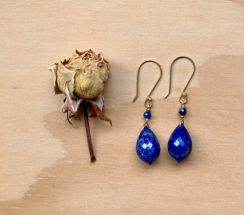 May include: A pair of gold earrings with teardrop-shaped lapis lazuli stones. The stones are a deep blue color and have a faceted finish. The earrings are suspended from gold hooks.