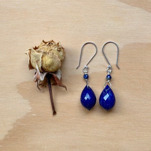 May include: A pair of silver earrings with teardrop-shaped lapis lazuli stones. The stones are a deep blue color and have a faceted finish. The earrings are hanging from silver hooks.