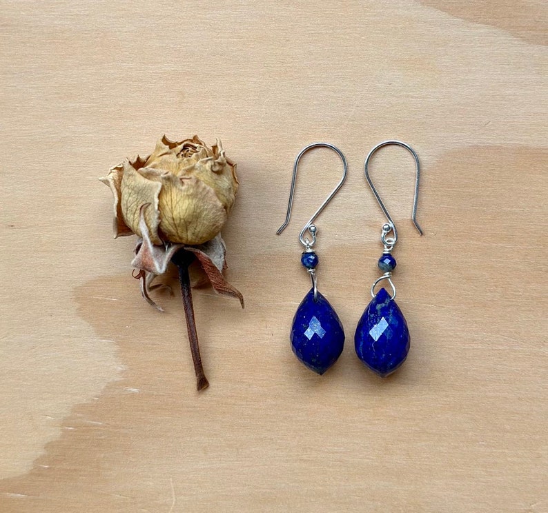 May include: A pair of silver earrings with teardrop-shaped lapis lazuli stones. The stones are a deep blue color and have a faceted finish. The earrings are hanging from silver hooks.