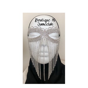 May include: A silver rhinestone mask with a fringe of rhinestones hanging from the bottom. The mask is on a white mannequin head with black fabric around the neck. The text "Boutique Al Jameelah" is written above the mask.