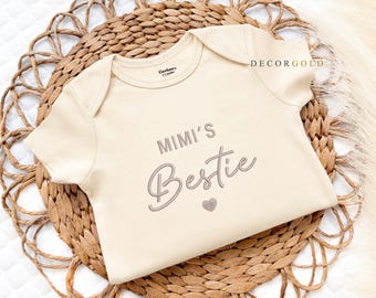 May include: A cream-colored baby onesie with short sleeves. The text "MIMI'S Bestie" is printed in gray script, with a small heart below. The onesie is placed on a woven, circular placemat. The brand name "DECORGOLD" is printed on the onesie.