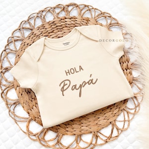 May include: A beige baby onesie with the words "HOLA Papá" in brown script. The onesie is made of soft material and is displayed on a woven, circular placemat. The brand name "DECORGOLD" is visible.