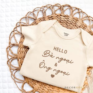 May include: A cream-colored baby onesie with the text "HELLO Bà ngoại & Ông ngoại" in brown script. The onesie is placed on a woven, circular placemat. The background is a white, quilted surface.