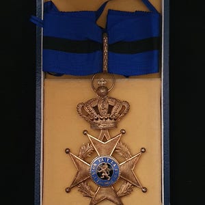 Pode incluir: Medalha dourada com design de cruz, com um emblema azul e branco no centro. A medalha é adornada com uma coroa e suspensa por uma fita azul com uma faixa preta, apresentada numa caixa azul com interior bege.