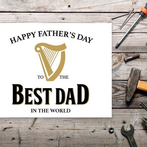 May include: White card with black text that reads "Happy Father's Day to the Best Dad in the World". A gold harp logo is above the text. The card is on a wooden surface with tools and nails.