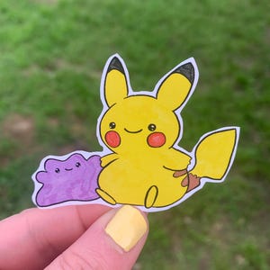 May include: A yellow Pikachu sticker with black-tipped ears, red cheeks, and a smiling face. A purple Ditto sticker is next to Pikachu. The stickers are held against a blurred green background.