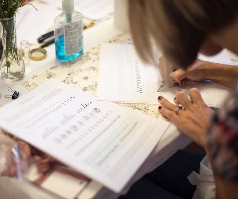 Puede incluir: Una persona practicando caligraf&iacute;a con una pluma y tinta sobre una hoja de papel blanca. El papel tiene l&iacute;neas y una secci&oacute;n con las palabras "ultra blue ultra" y una serie de c&iacute;rculos.