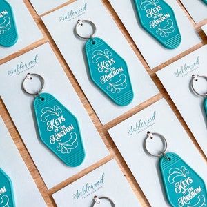 K&ouml;nnte beinhalten: T&uuml;rkise Motel-Schl&uuml;sselanh&auml;nger mit wei&szlig;er Aufschrift "Keys to the Kingdom". Die Schl&uuml;sselanh&auml;nger werden auf wei&szlig;en Karten mit dem Logo der Sablewood Paper Company pr&auml;sentiert.