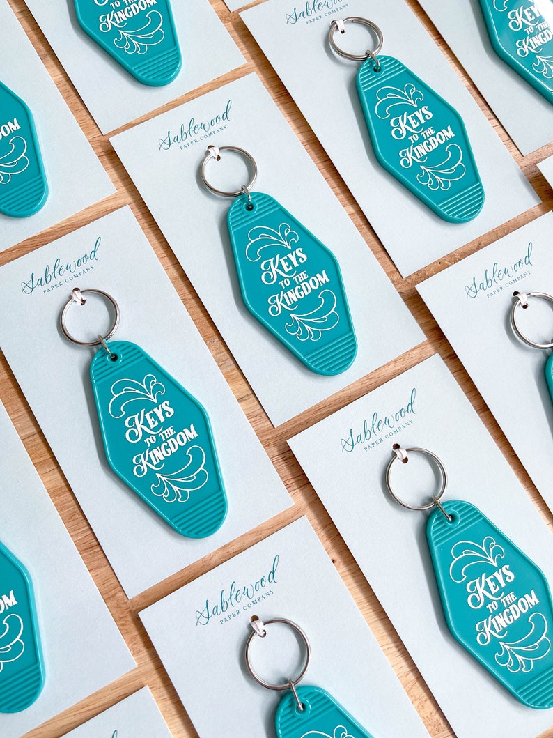 K&ouml;nnte beinhalten: T&uuml;rkise Motel-Schl&uuml;sselanh&auml;nger mit wei&szlig;er Aufschrift "Keys to the Kingdom". Die Schl&uuml;sselanh&auml;nger werden auf wei&szlig;en Karten mit dem Logo der Sablewood Paper Company pr&auml;sentiert.