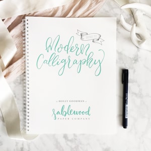 Puede incluir: Una libreta blanca de espiral con el t&iacute;tulo "Intro to Modern Calligraphy" escrito en cursiva turquesa. La libreta tambi&eacute;n presenta un logotipo turquesa que dice "Sablewood Paper Company" con un adorno decorativo.