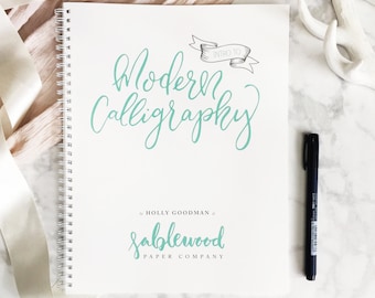 May include: A white spiral bound notebook with the title "Intro to Modern Calligraphy" written in teal cursive. The notebook also features a teal logo that says "Sablewood Paper Company" with a decorative flourish.