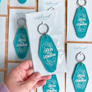 K&ouml;nnte beinhalten: T&uuml;rkise Motel-Schl&uuml;sselanh&auml;nger mit wei&szlig;er Aufschrift "Keys to the Kingdom". Die Schl&uuml;sselanh&auml;nger sind in durchsichtigen Plastikt&uuml;ten verpackt, mit einer wei&szlig;en Karte, auf der "Sablewood Paper Company" steht.