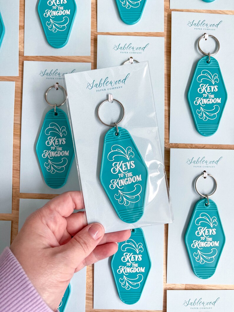K&ouml;nnte beinhalten: T&uuml;rkise Motel-Schl&uuml;sselanh&auml;nger mit wei&szlig;er Aufschrift "Keys to the Kingdom". Die Schl&uuml;sselanh&auml;nger sind in durchsichtigen Plastikt&uuml;ten verpackt, mit einer wei&szlig;en Karte, auf der "Sablewood Paper Company" steht.
