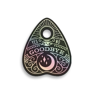 May include: A black and white Ouija board planchette with a holographic finish. The planchette is shaped like a teardrop and has a hole in the top. The words "YES" and "NO" are printed on the planchette, along with the word "GOODBYE" in a decorative font.