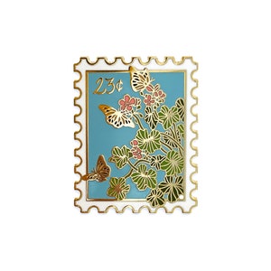 May include: A gold-tone enamel pin shaped like a postage stamp with a blue background. The stamp features a floral design with pink flowers and green leaves, and two butterflies. The stamp also has the text "23¢" printed on it.