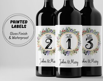 May include: Three wine bottles with labels featuring a watercolor wreath design with gold accents. The labels include the text "Table" and a number, "2", "1", and "3". The labels also include the text "John & Mary".