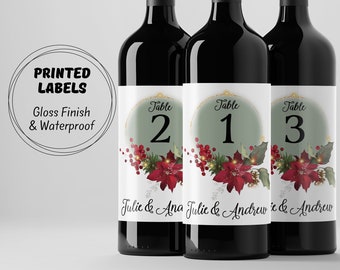 May include: Three wine bottles with Christmas-themed labels. The labels are green and white with gold accents and feature a poinsettia flower, holly, and berries. The labels also include the text "Table" and a table number, as well as the names "Julie & Ana", "Julie & Andrew", and "e & Andrew".