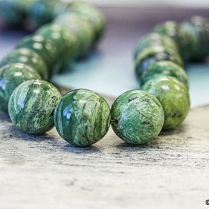 May include: A strand of green serpentine beads, each bead is round and has a unique marbled pattern.