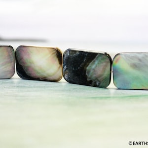 May include: A close-up of a row of iridescent rectangular shell beads. The beads are a mix of white, black, and green.