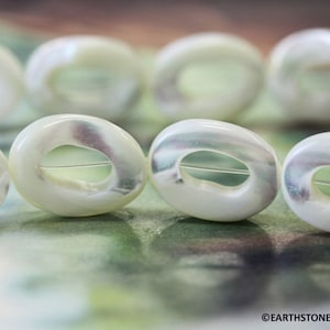 May include: A strand of white oval-shaped shell beads with a hole in the center of each bead. The beads have a slight iridescent sheen.