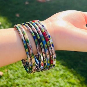 May include: A set of colourful enamel bangles. The bangles have a floral design with red, blue, green, and white accents. The bracelets are stacked on a wrist against a blurred green background.