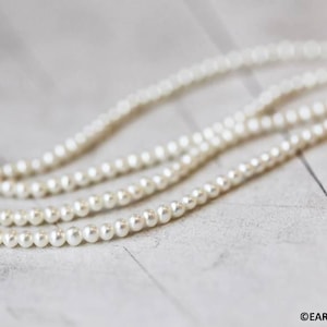 May include: A strand of white pearls, arranged in a loose, flowing pattern on a light gray surface.