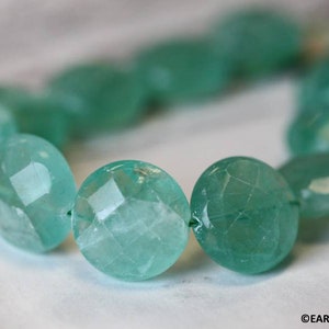 May include: A strand of faceted, light green gemstone beads. The beads are round and have a slightly irregular shape. The beads are strung on a thin, clear thread.