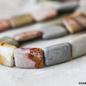May include: Close-up of a strand of square-shaped, multi-colored gemstone beads. The beads are a mix of light brown, pink, gray, and white.