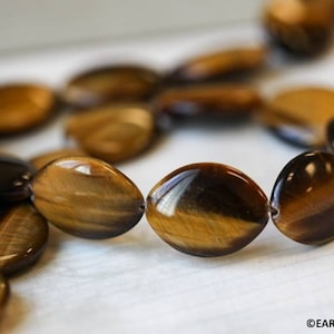 May include: A strand of oval-shaped tiger's eye beads in shades of brown and gold. The beads are polished and have a glossy finish.