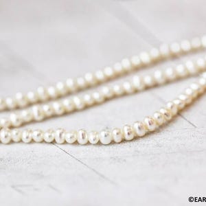 May include: A strand of white freshwater pearls, strung on a thin thread, lies on a white surface. The pearls are small and round, with a smooth, glossy finish.