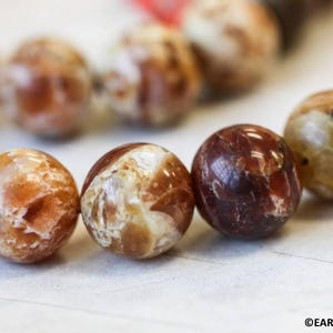 May include: Close-up of a strand of polished, round gemstone beads. The beads are a mix of brown, tan, and cream colors with natural patterns. The beads are strung together, suggesting a necklace or bracelet.