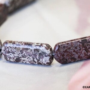 May include: A close-up of a necklace made of brown and white speckled stone beads. The beads are rectangular in shape and have a polished finish.