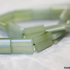 May include: Close-up of a strand of light green jade beads. The beads are rectangular with slightly rounded edges and are strung together with a green thread. The beads have a translucent quality and a subtle sheen.