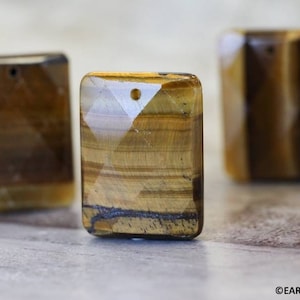 May include: Three rectangular tiger's eye gemstone pendants with a hole drilled through the top for stringing. The stones are a mix of brown, gold, and black.