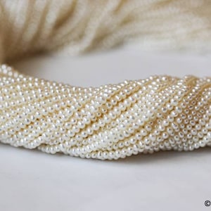 May include: A close-up of a strand of white freshwater pearls. The pearls are small and round, and they are strung together on a thin thread. The pearls are arranged in a loose, flowing pattern.