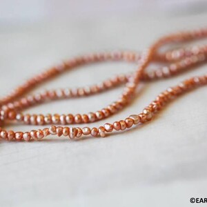 May include: A strand of small, irregular, copper-colored pearls with a lustrous finish. The pearls are strung together in a continuous line, creating a textured appearance. The text EARTHSTONE.COM is visible in the bottom right corner.
