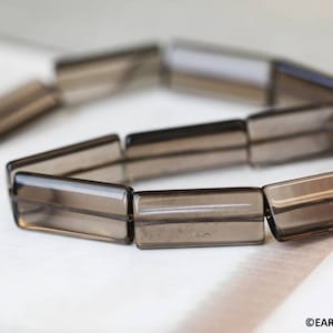 May include: A strand of smoky quartz beads, each bead is a rectangular prism with a smooth, polished finish. The beads are strung together on a clear thread.