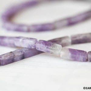 May include: Close-up of a strand of amethyst beads. The beads are rectangular with a mix of purple and white hues. The beads are strung together, with some in focus and others blurred in the background. The image is on a white surface.