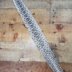 May include: A long, narrow strip of snakeskin featuring a repeating diamond pattern in shades of gray, black, and white. The texture is scaled and detailed, displaying a natural, organic design that runs the entire length of the elongated piece.