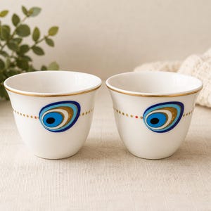 May include: Two white ceramic cups featuring a gold rim and a central eye-shaped motif in shades of blue, gold, and black. A horizontal line of small gold dots encircles the exterior of each cup, accentuating the central graphic design.