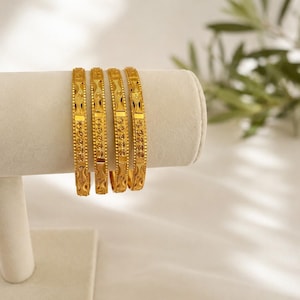 May include: A set of four gold bangles featuring a repeating textured pattern with small raised dots and vertical rectangular segments. Each bangle has a polished metallic finish and a consistent width, displaying intricate geometric detailing.