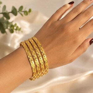May include: A set of four gold-toned bangles featuring a repeating embossed pattern of small circles and elongated oval shapes. The bracelets have a polished, reflective finish and are worn together on the wrist, creating a stacked appearance.